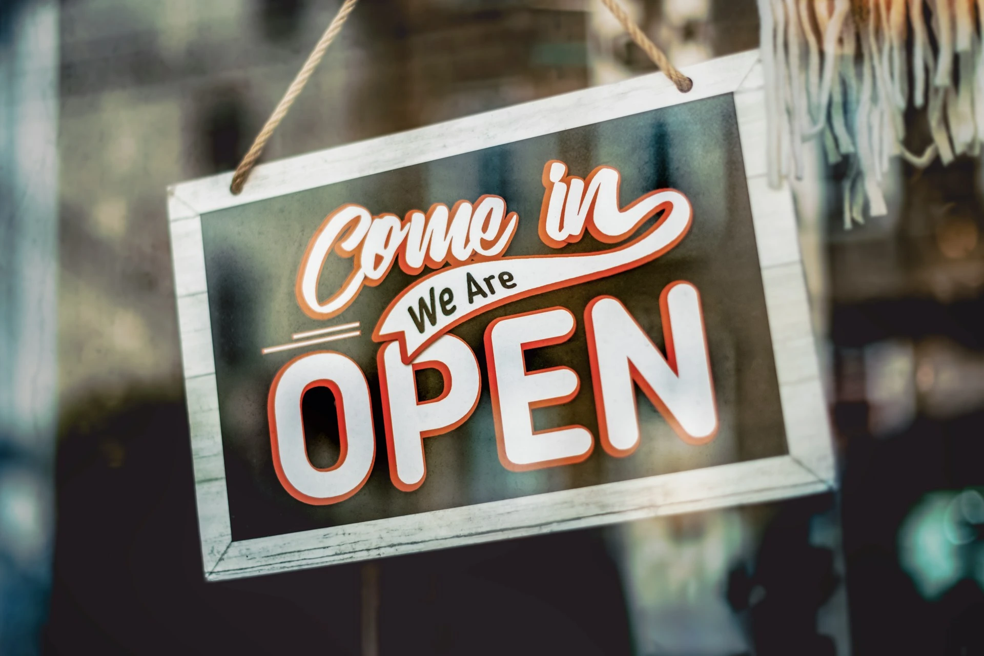 A neon sign in a window reads "come in we are open," hanging from a metal chain with a blurred background, inviting the brand community inside.