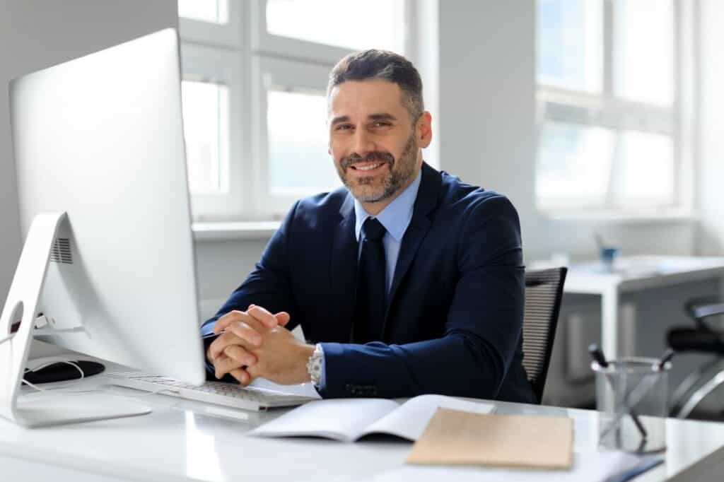 A man wearing a suit and tie sits at a desk with a computer monitor, keyboard, and open documents in a well-lit office. Smiling and looking directly at the camera, he embodies the professionalism of DigiSmart, a leading digital marketing agency.