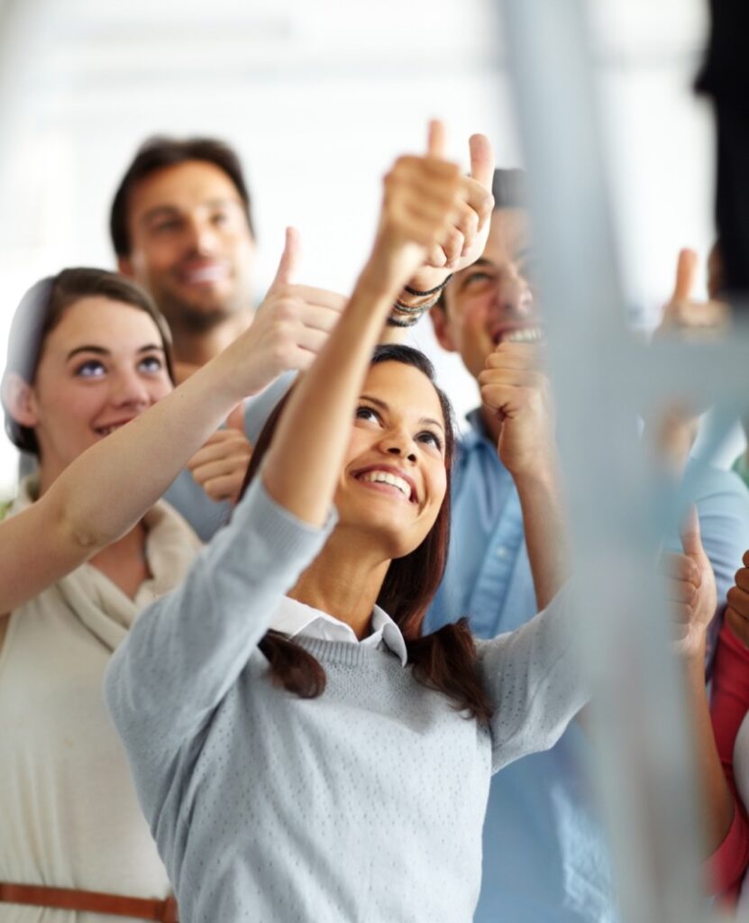 A group of people stand close together indoors, smiling and giving thumbs-up gestures, appearing positive and supportive.